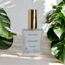 Load image into Gallery viewer, This image features one elegantly designed bottle of Eau De Parfum,with gold cap. and are presented on a wooden surface with large green leaves in the background, enhancing the natural and sophisticated appeal of the products.
