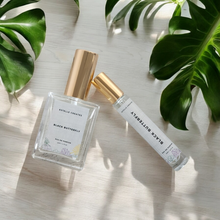 Load image into Gallery viewer, This image features two elegantly designed bottles of Eau De Parfum, one larger and one smaller, both with gold caps. and are presented on a wooden surface with large green leaves in the background, enhancing the natural and sophisticated appeal of the products.

