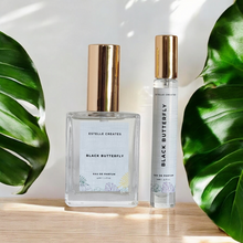 Load image into Gallery viewer, This image features two elegantly designed bottles of Eau De Parfum, one larger and one smaller, both with gold caps. The bottles are labeled "Black Butterfly" and are presented on a wooden surface with large green leaves in the background, enhancing the natural and sophisticated appeal of the products.