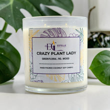 Load image into Gallery viewer, Candle labeled 'Crazy Plant Lady' with a decorative label on a white surface with green leaves in the background. Green floral, fig, wood