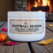 Load image into Gallery viewer, Candle labeled 'Football Season' on a wooden surface with a fireplace in the background