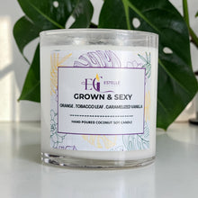 Load image into Gallery viewer, Grown and SexyCandle with a label on a white surface with green leaves in the background. Orange tobacco leaf caramelized vanilla