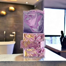 Load image into Gallery viewer, 4 soap bars stacked in a bathroom setting. purple soap. Indulge in our  handmade artisan soap bar that transforms your daily routine into a luxurious escape.