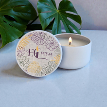 Load image into Gallery viewer, Candle with decorative lid featuring 'Estelle Creates' branding on a light surface with green leaves in the background.