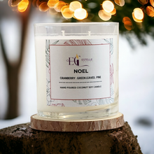 Load image into Gallery viewer, Candle labeled 'Estelle Creates' with 'NOEL' scent on a wooden coaster against a blurred festive background.