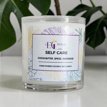 Load image into Gallery viewer, Self care Candle with a label on a white surface with self green leaves in the background. Cocoa butter spices olive wood